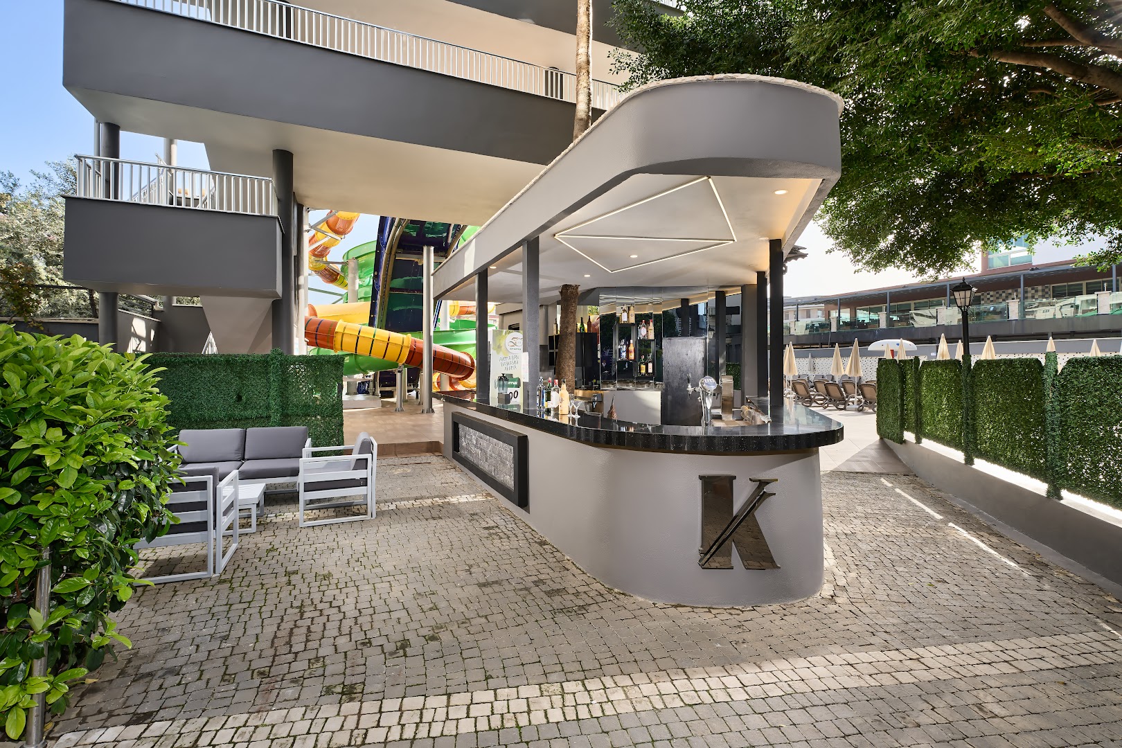 Kaila Beach Hotel āra bārs ar modernu dizainu un atpūtas zonu — fonā redzami krāsaini ūdens slidkalniņi un sauļošanās krēsli