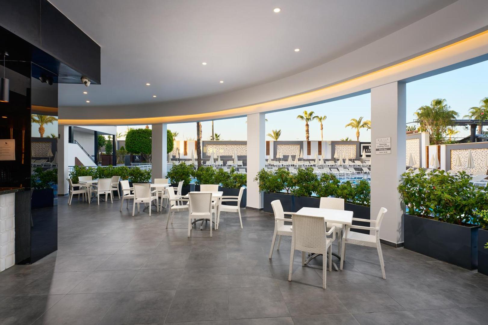 Kaila Beach Hotel āra terases bārs ar baltām mēbelēm un skatu uz baseina zonu un palmām saulrieta gaismā