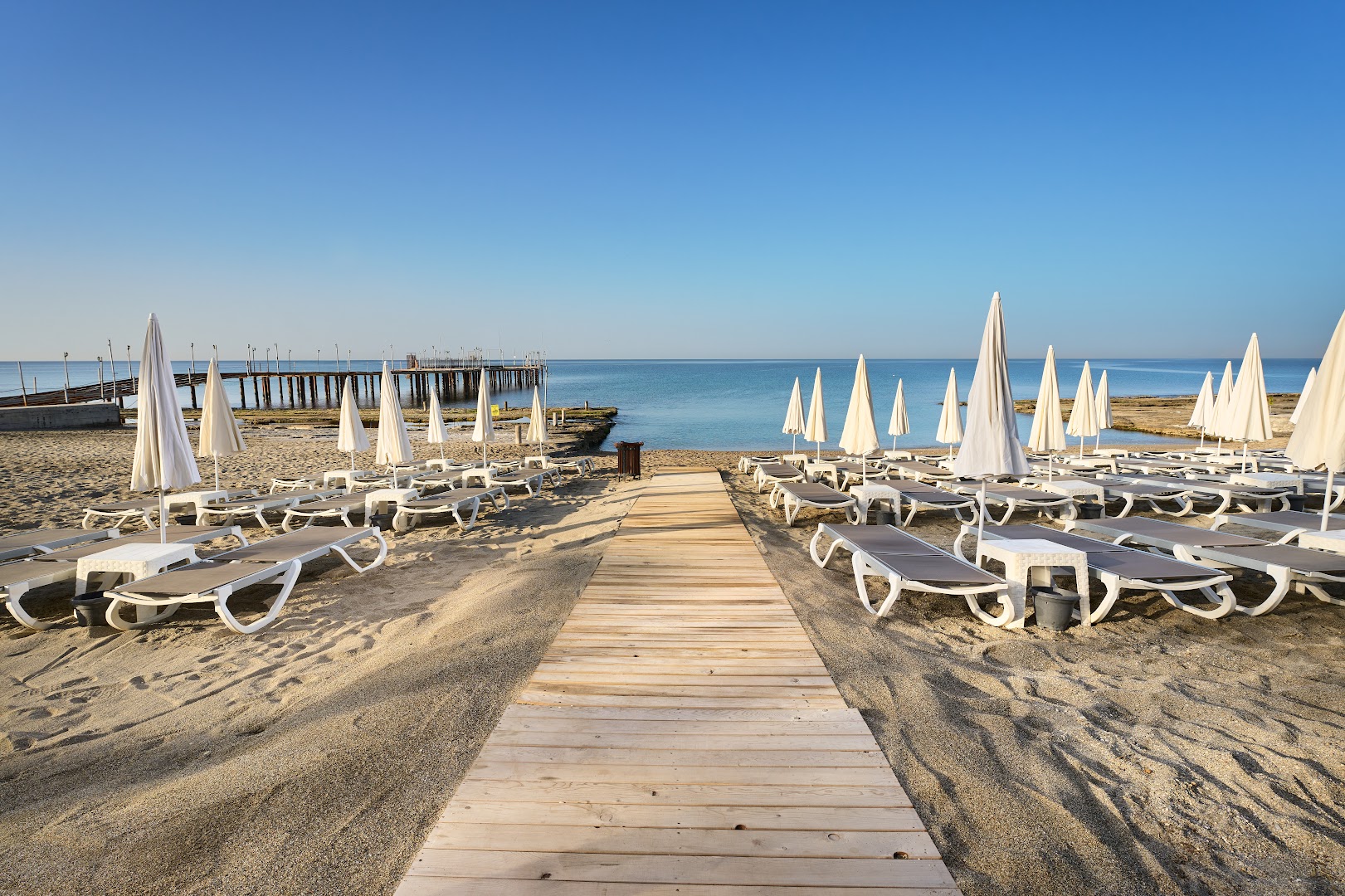 Kaila Beach Hotel koka laipa uz pludmali ar sauļošanās krēsliem un saulessargiem abās pusēs, fonā jūra un mols