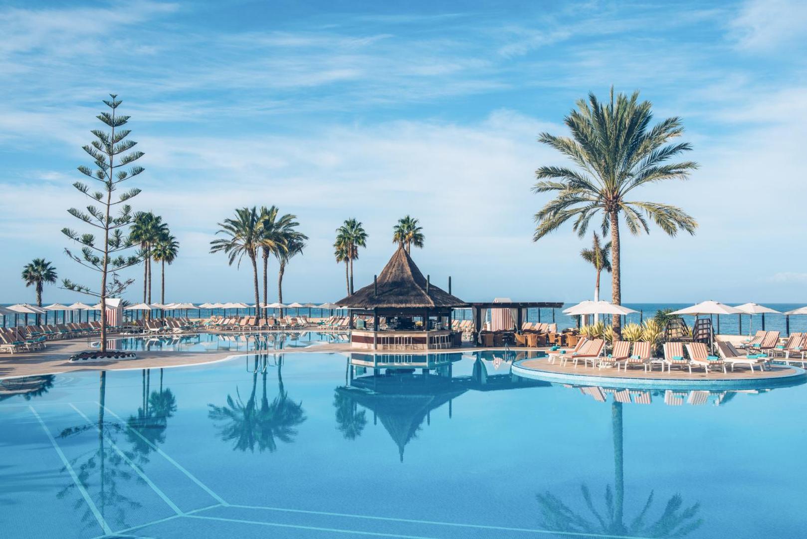 Iberostar Selection Anthelia bārs baseina vidū ar salmu jumtu Tenerifē — apkārt sauļošanās zona ar krēsliem un skats uz okeānu.