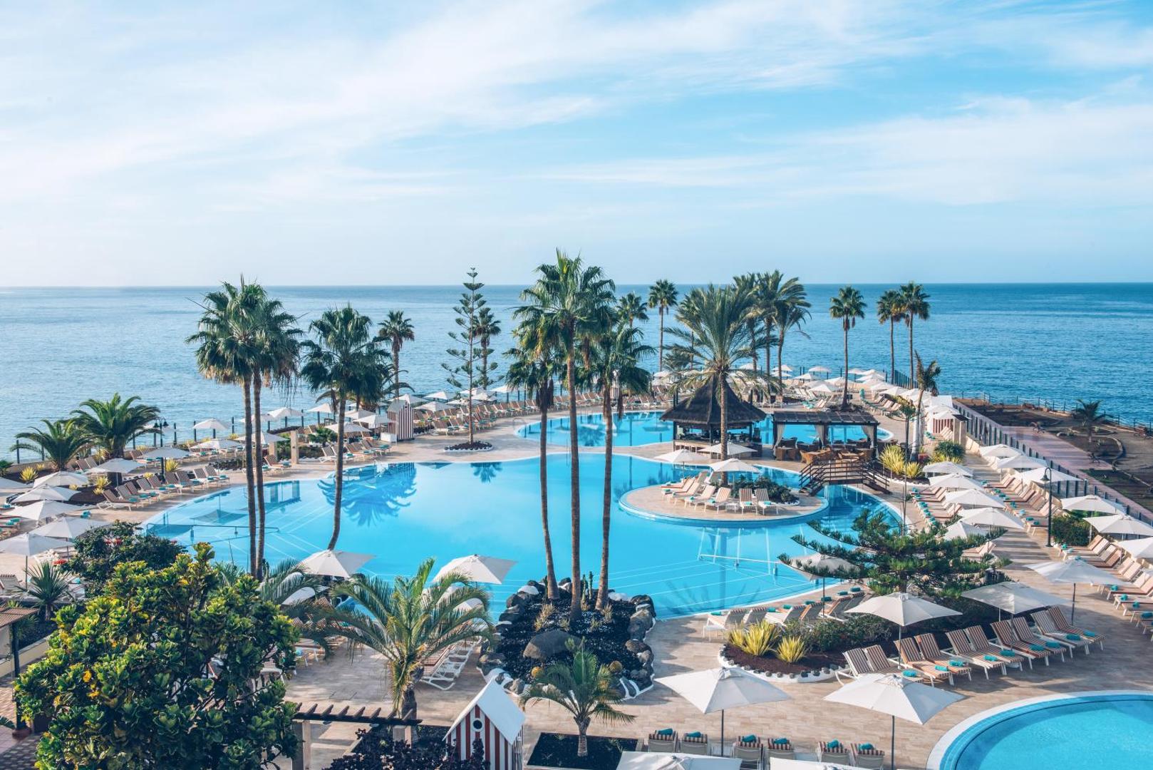 Iberostar Selection Anthelia plašais āra baseinu komplekss ar palmām un skatu uz okeānu Tenerifē — sauļošanās krēsli un saulessargi pie ūdens