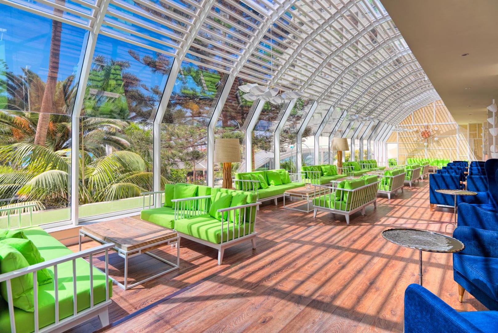 H10 Las Palmeras gaišā terase ar stikla sienām, zaļiem dīvāniem un skatu uz palmām piekrastē Tenerifē