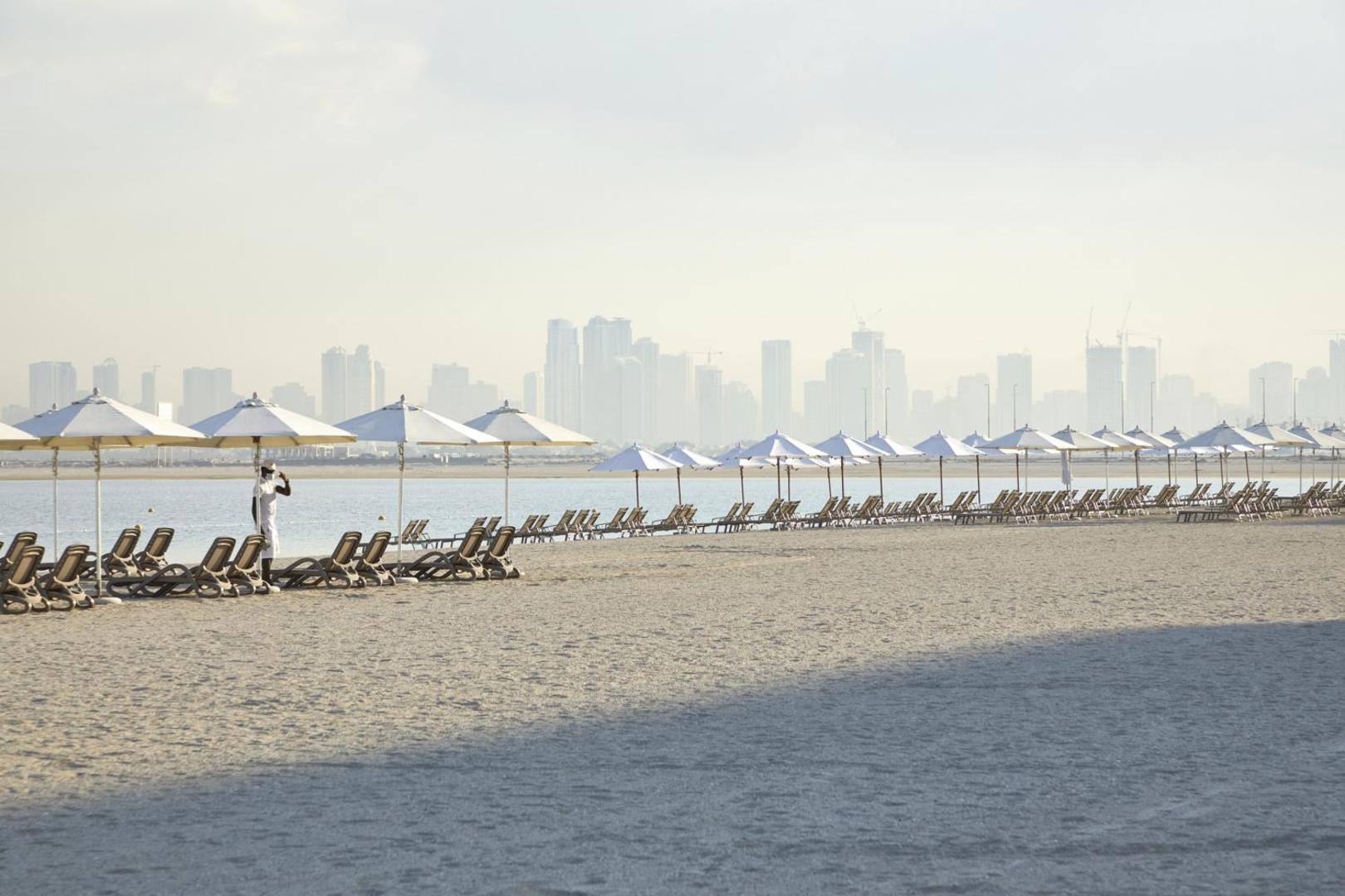 RIU Dubai privātā smilšu pludmale ar gariem sauļošanās krēslu un saulessargu rindām, fonā redzama Dubaijas pilsētas panorāma.