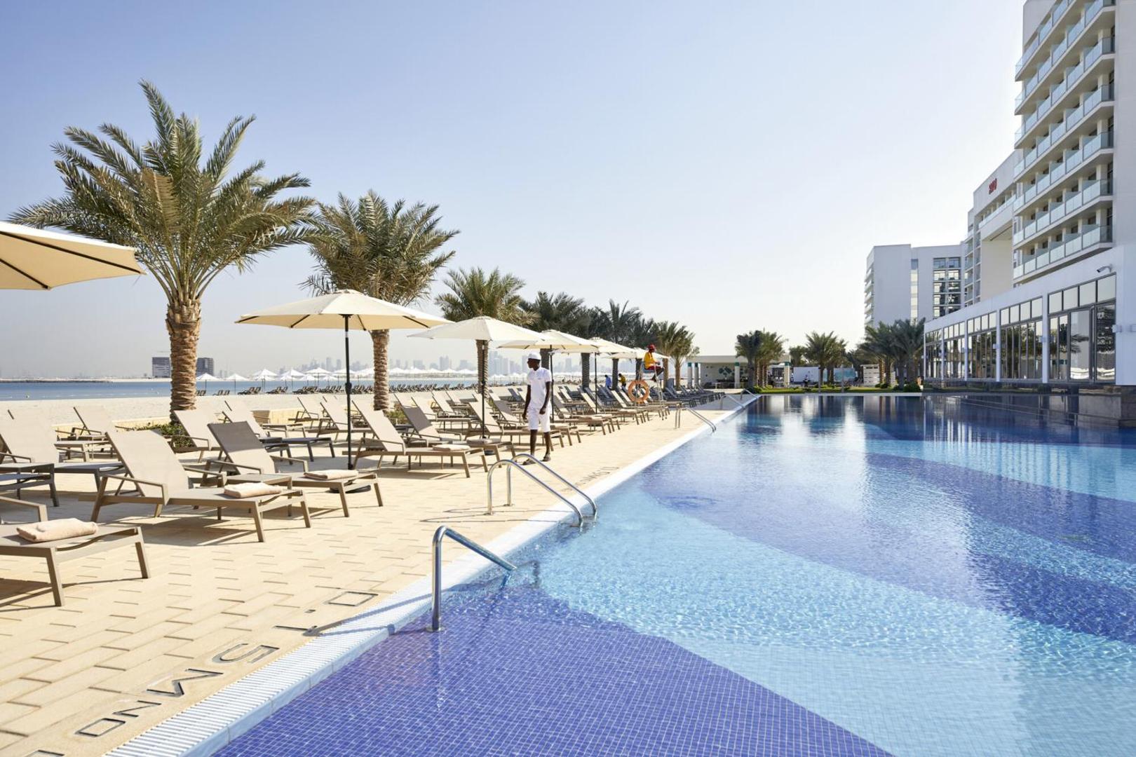 RIU Dubai āra baseins ar zilu flīžu apdari, sauļošanās krēsliem un saulessargiem — blakus pludmale un palmas