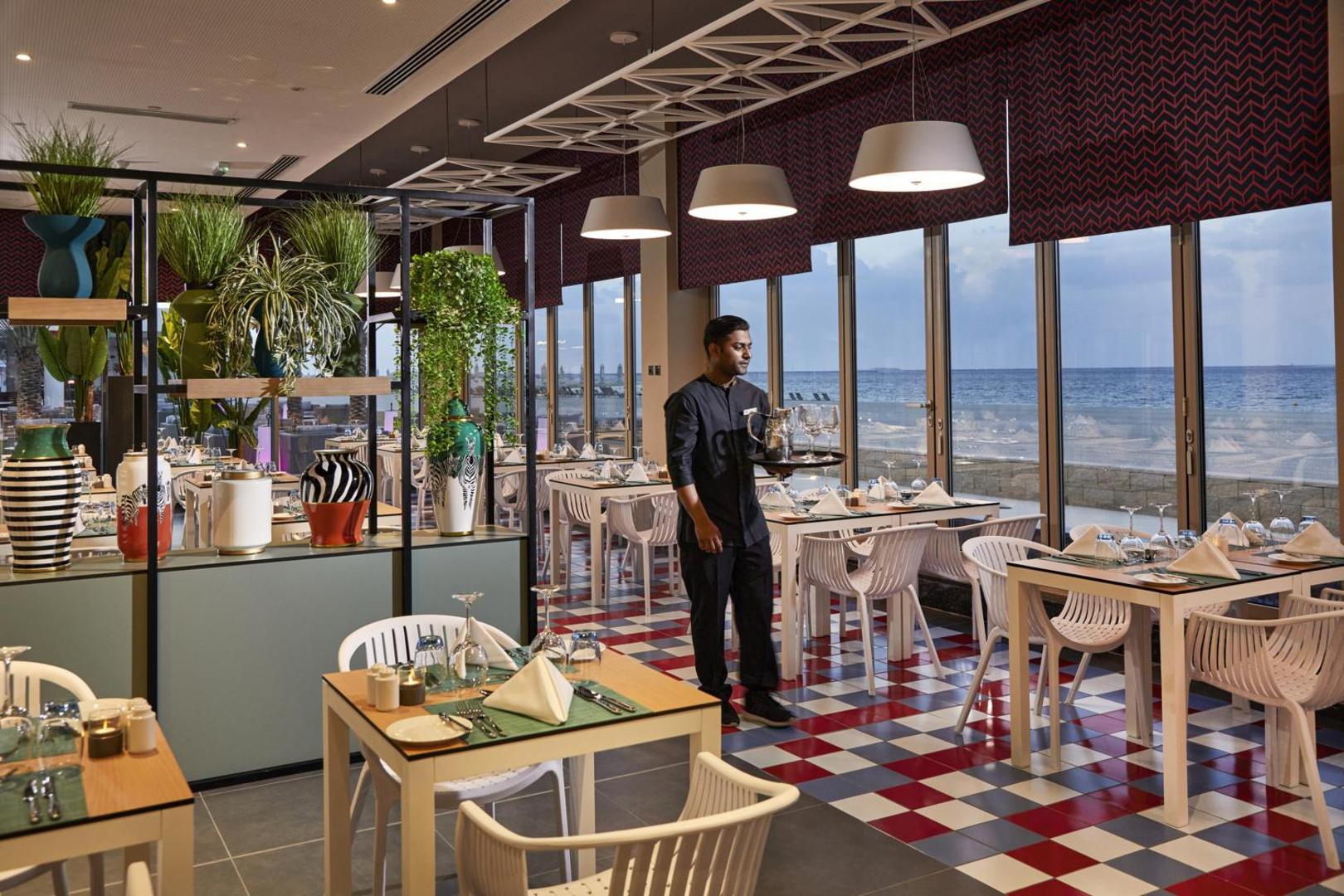 RIU Dubai viesnīcas restorāns ar skatu uz jūru, modernu interjeru, dekoratīviem augiem un viesmīli, kas pasniedz dzērienus pie galdiņiem.