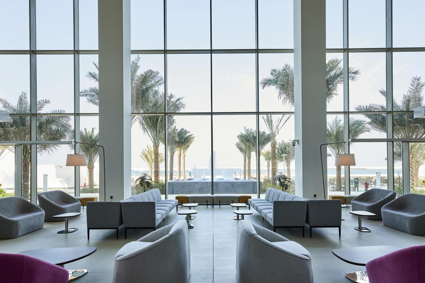 Atpūtas zona RIU Dubai viesnīcā ar mīkstajiem krēsliem pie panorāmas logiem, aiz kuriem redzamas palmas un jūra