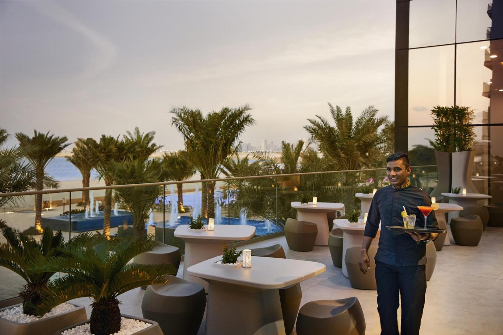 RIU Dubai āra terase ar skatu uz Persijas līci un palmām saulrietā, viesmīlis pasniedz eksotiskus kokteiļus