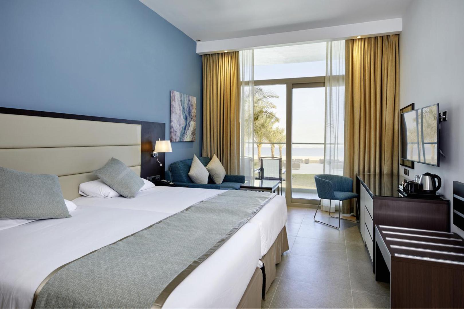 RIU Dubai gaišs divvietīgs numurs ar balkonu, no kura paveras skats uz palmām un jūru — zilas sienas un mēbeles