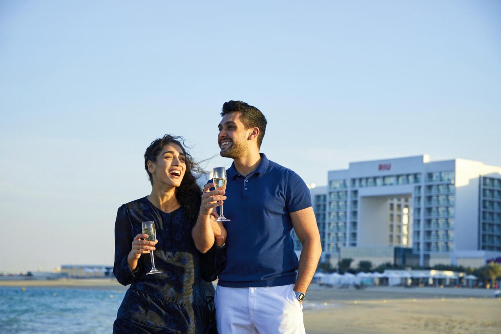 Laimīgs pāris ar dzērienu glāzēm RIU Dubai pludmalē uz viesnīcas ēkas fona saulrieta laikā