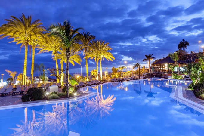 Melia Jardines del Teide āra baseins vakarā ar izgaismotām palmām un zilu ūdeni piekrastē, Tenerifē, Kanāriju salās.