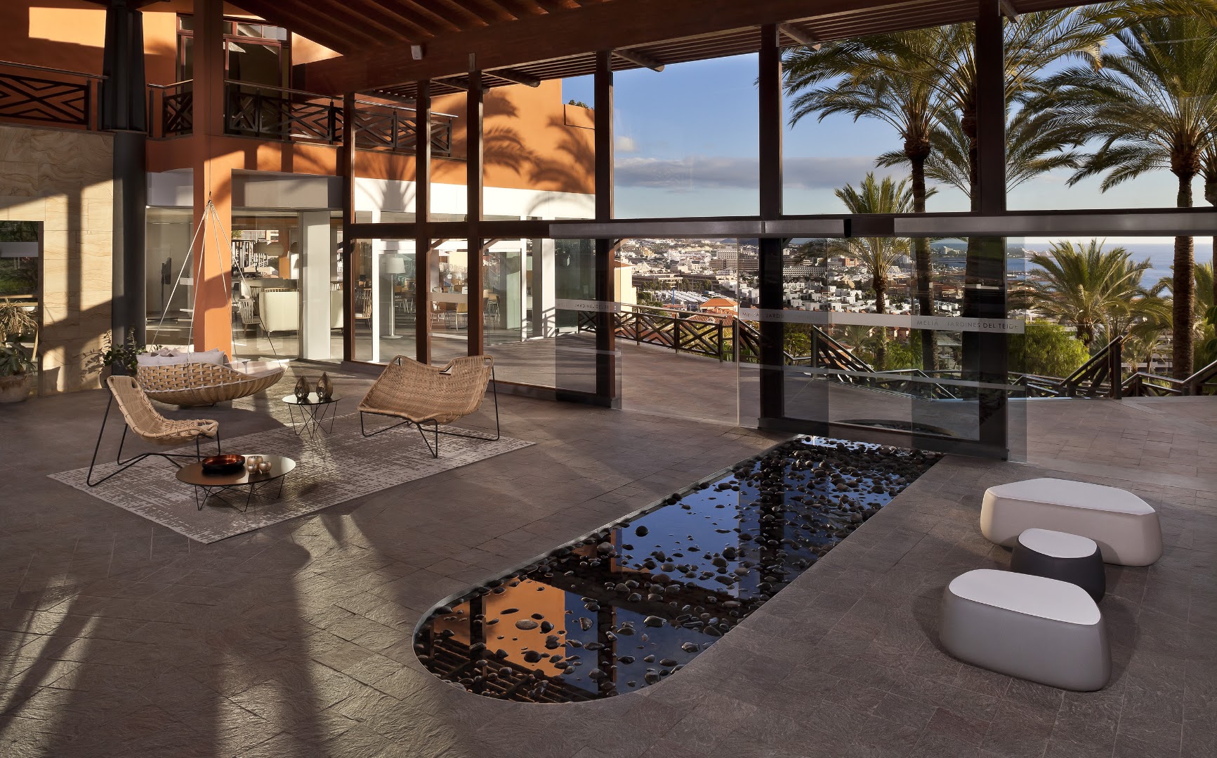 Melia Jardines del Teide viesnīcas lobby atpūtas zona ar stilīgām mēbelēm, dekoratīvu ūdens elementu un panorāmas skatu uz pilsētu.