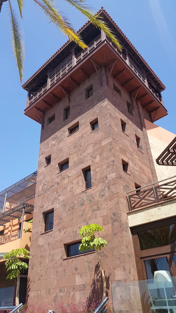 Meliá Jardines del Teide raksturīgais sarkana akmens tornis ar koka balkonu pret zilām debesīm un palmas lapām