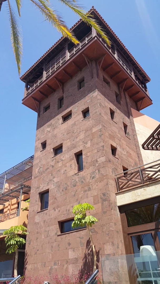 Melia Jardines del Teide arhitektūras elements — akmens mūra tornis ar koka balkonu un maziem logiem pret zilu debesu fonu