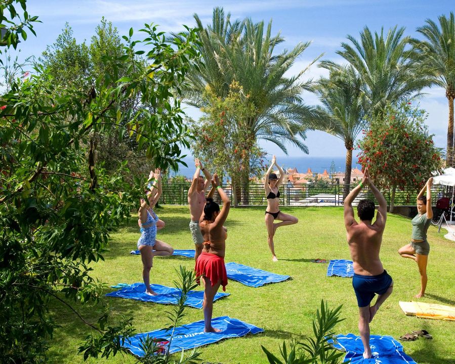 Viesu grupa nodarbojas ar jogu Melia Jardines del Teide zaļajā dārzā palmu ieskautā zālienā ar skatu uz jūru.
