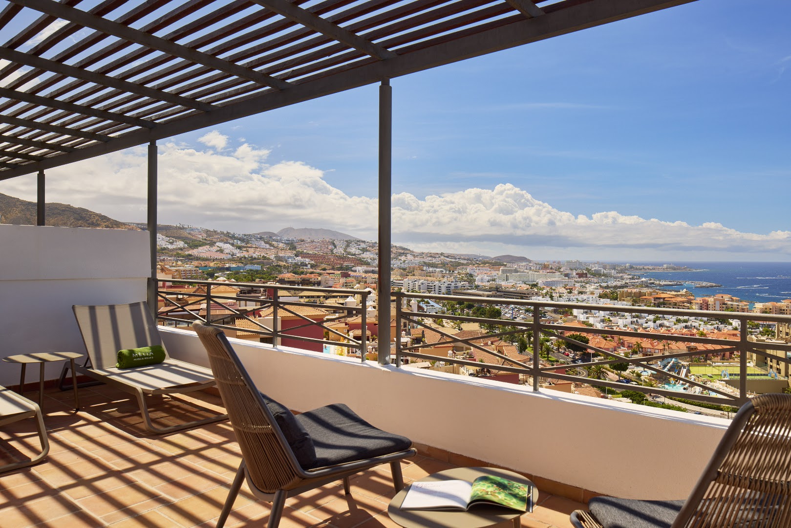 Skats no Melia Jardines del Teide terases uz kūrortpilsētu un piekrasti — atpūtas krēsli zem koka nojumes saulainā dienā