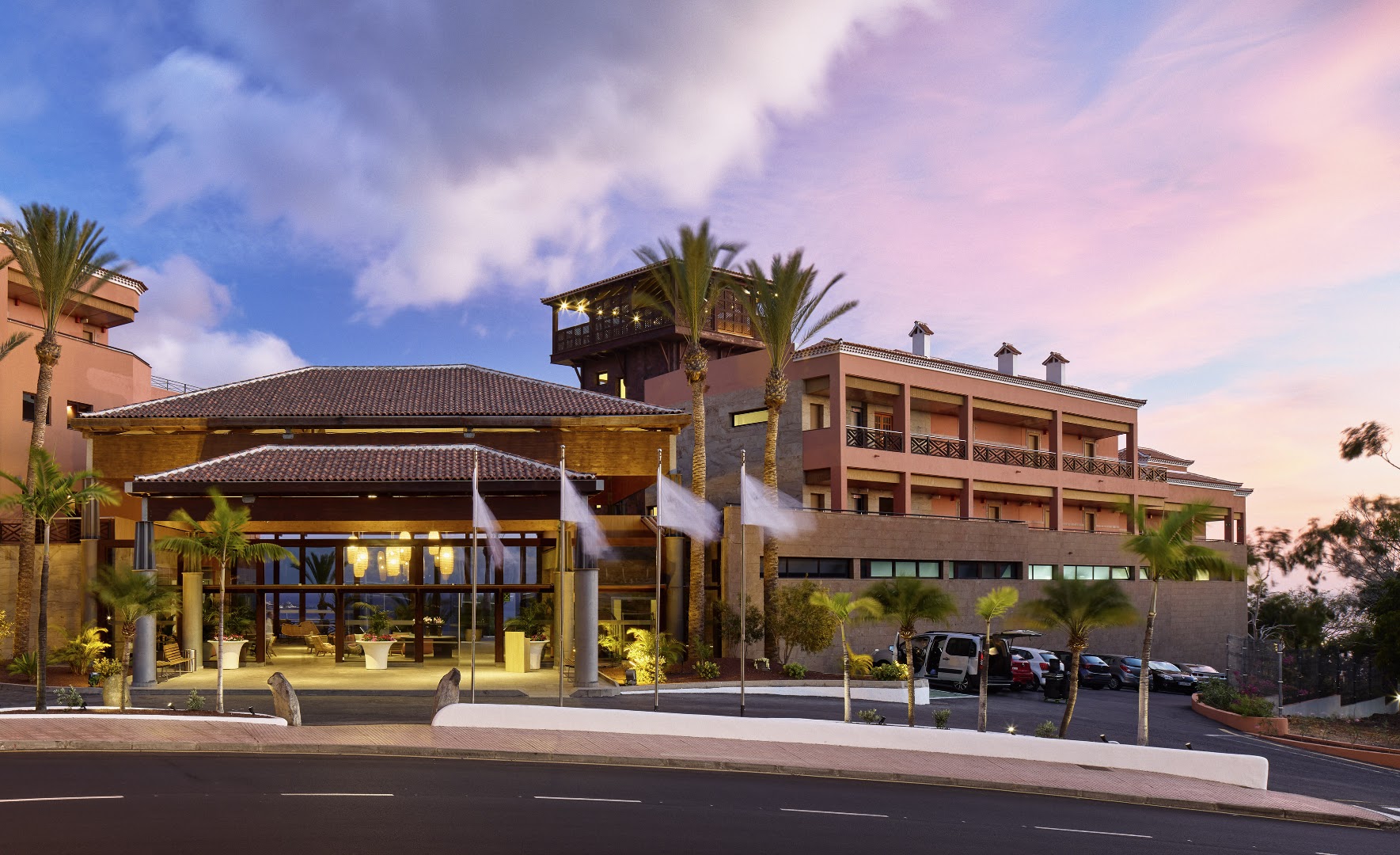 Melia Jardines del Teide viesnīcas galvenā ieeja un fasāde saulrieta laikā Tenerifē — apgaismota recepcijas zona ar palmām un karogiem