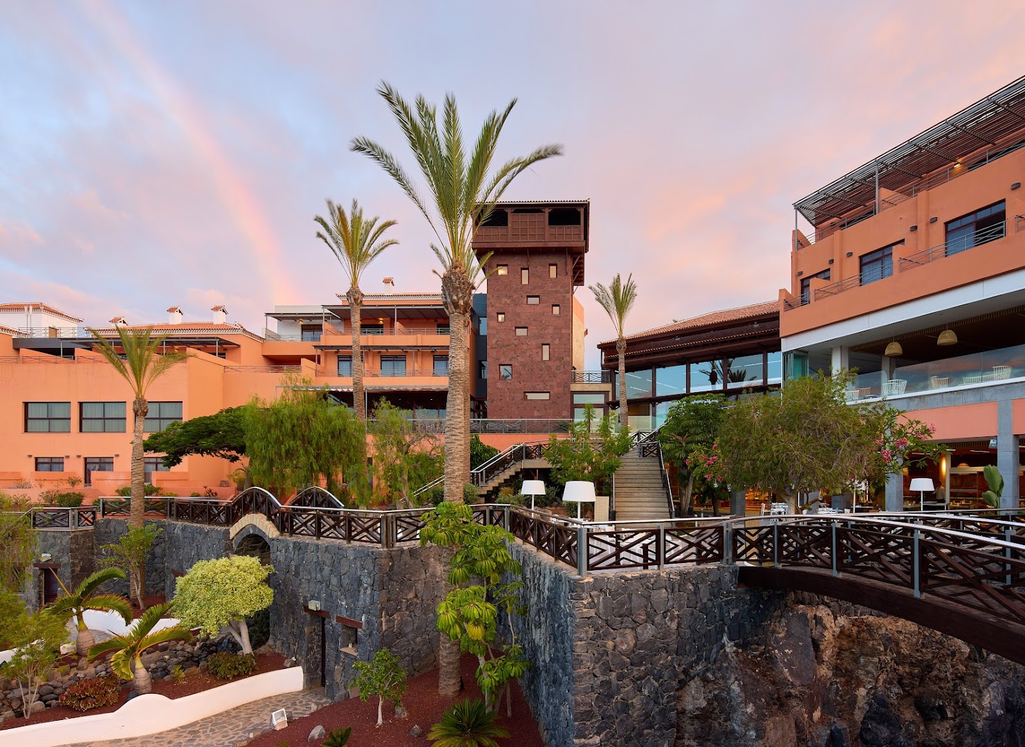 Meliá Jardines del Teide arhitektūra ar akmens sienām, koka tiltiņiem un terasēm uz rozā debesu un varavīksnes fona