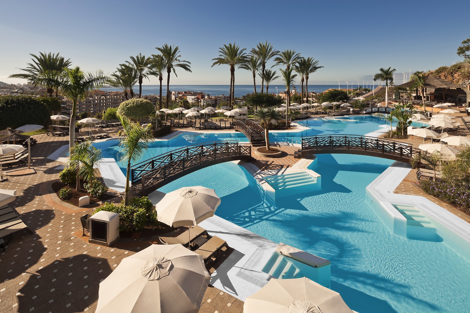 Melia Jardines del Teide baseinu zona ar dekoratīviem tiltiņiem un atpūtas krēsliem, fonā redzama pilsētas panorāma un okeāns