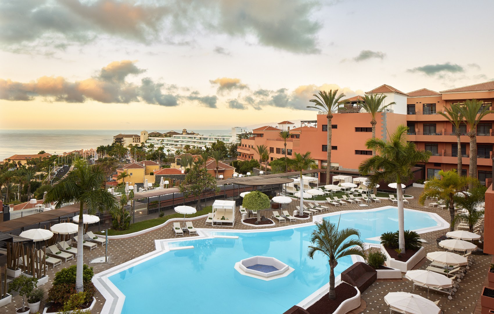 Melia Jardines del Teide viesnīcas ēkas arhitektūra saulrietā ar baseinu, atpūtas krēsliem un palmām piekrastes zonā.