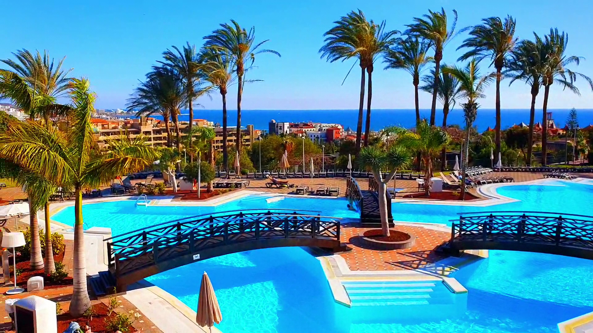Melia Jardines del Teide baseins ar koka tiltiņiem un palmām uz zila debesu un okeāna fona Tenerifē.