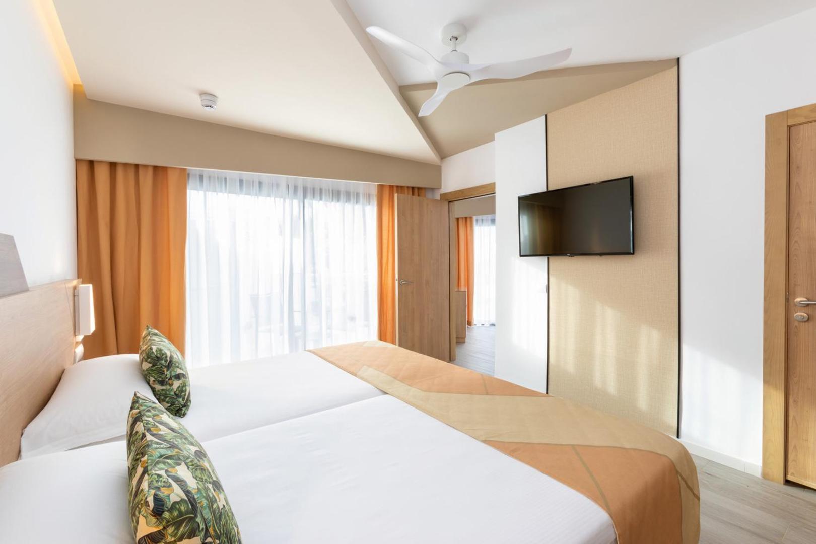 Riu Buena Vista numura skats no gultas puses uz logu un televizoru — gaiši aizkari un griestu ventilators komfortam