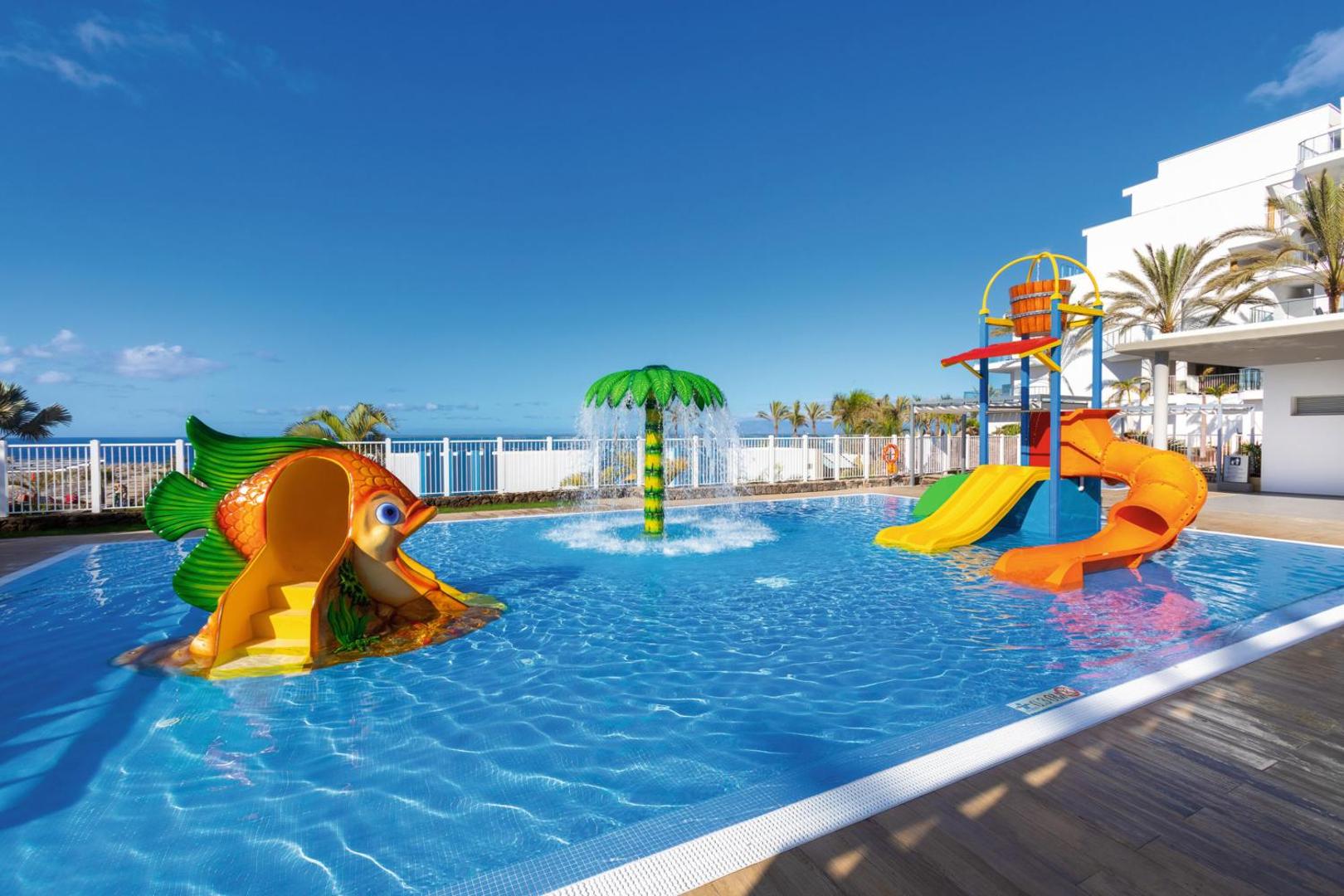 Riu Buena Vista bērnu ūdens atrakciju parks Tenerifē ar slidkalniņiem un palmas formas strūklaku uz viesnīcas korpusa fona
