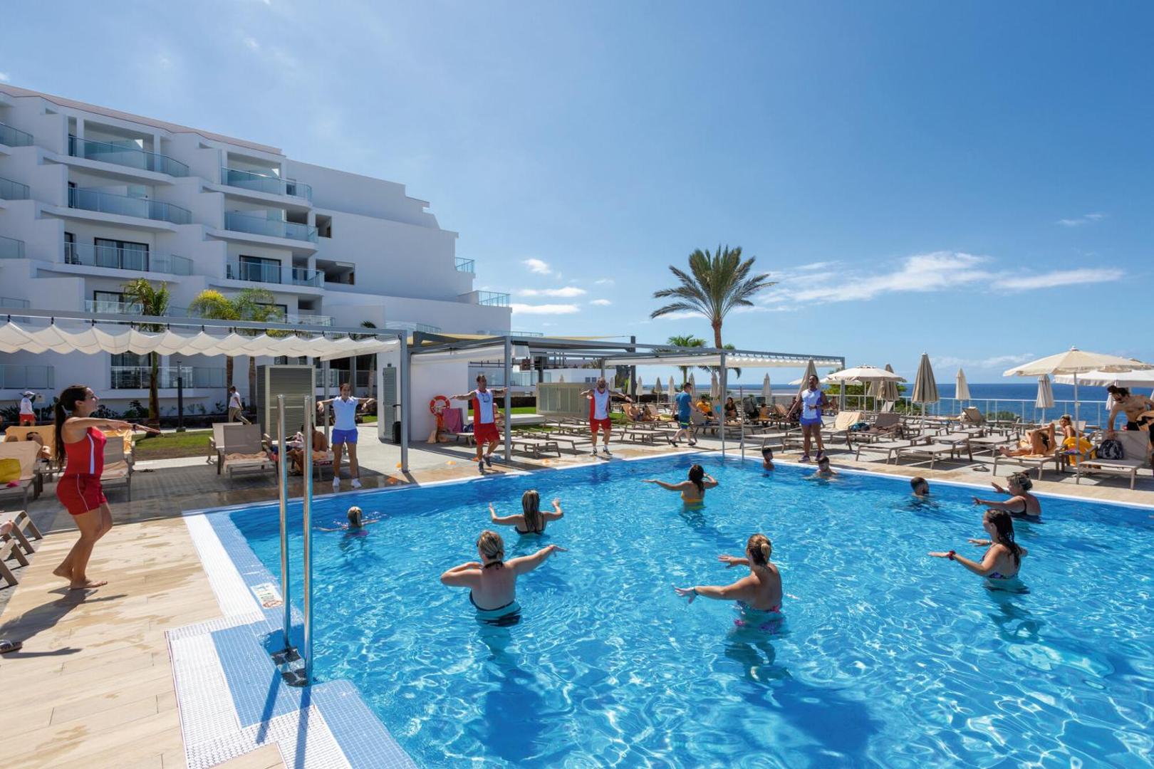 Riu Buena Vista āra baseins ar ūdens aerobikas nodarbību Tenerifē — viesi sporto saulainā terasē ar skatu uz okeānu