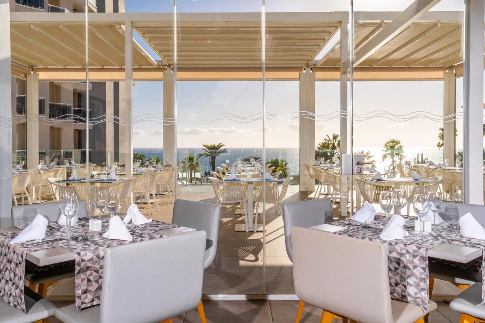 Hovima Cool Costa Adeje restorāna terase ar skatu uz Atlantijas okeānu un palmām saulrietā Tenerifē