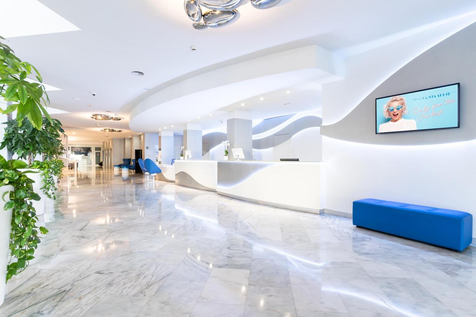 Hovima Cool Costa Adeje modernā viesnīcas lobby zona ar baltu recepcijas leti, marmora grīdu un stilīgu apgaismojumu Tenerifē