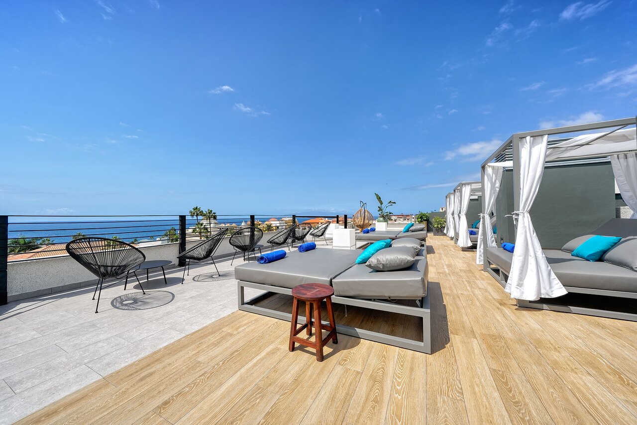 Hovima Cool Costa Adeje viesnīcas jumta terase Tenerifē ar moderniem atpūtas zviļņiem, saulessargiem un skatu uz okeānu saulainā dienā.
