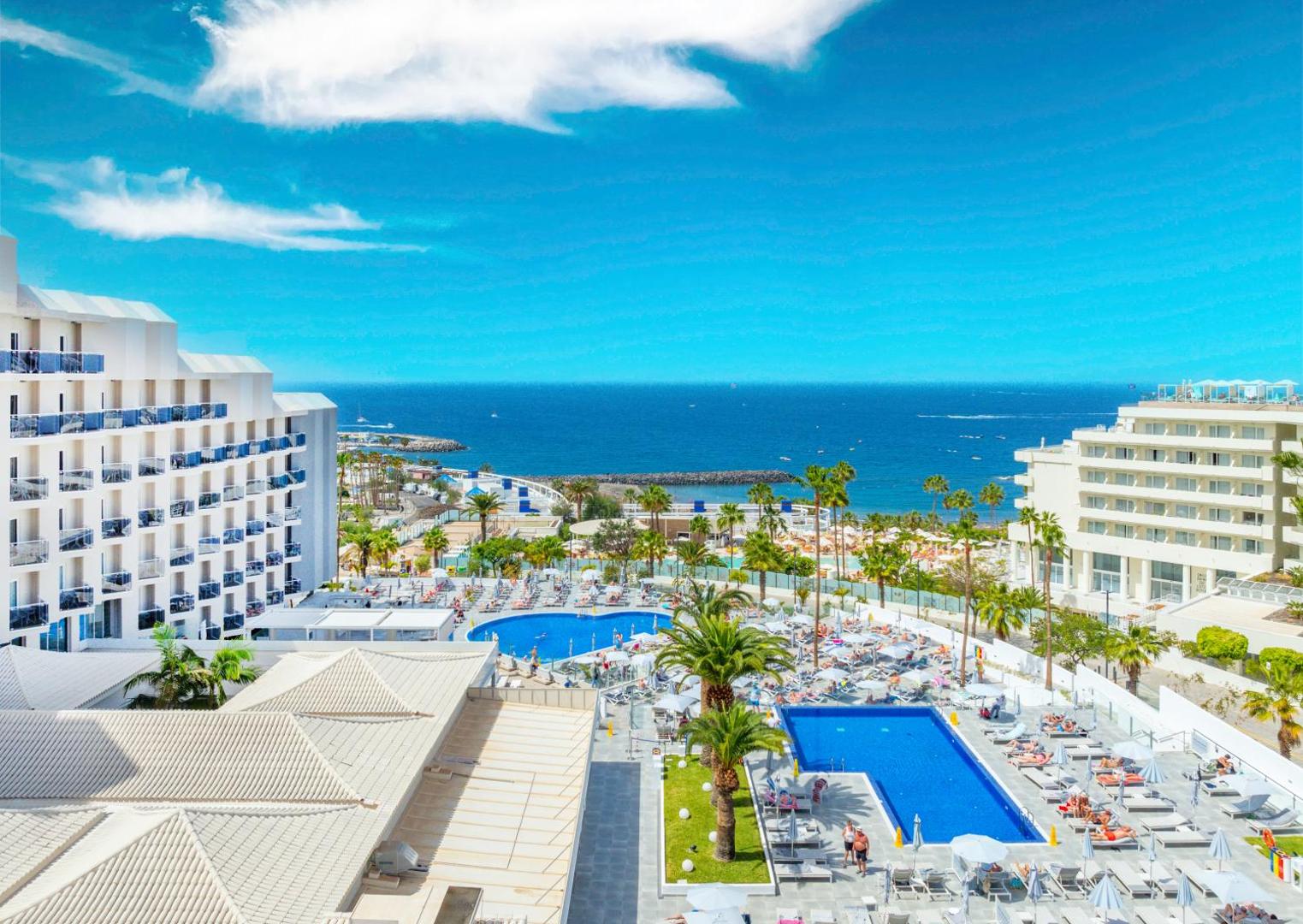 Hovima Cool Costa Adeje skats no augšas uz āra baseiniem un okeāna piekrasti Tenerifē — sauļošanās terase ar palmām un atpūtas krēsliem