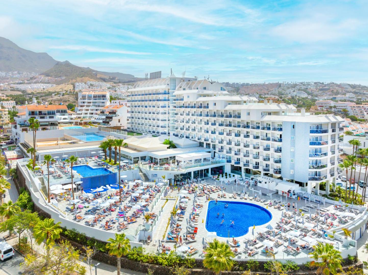 Hovima Cool Costa Adeje viesnīcas ēkas fasāde un plašā baseinu zona Tenerifē — panorāmas skats uz kūrortu un kalniem fonā
