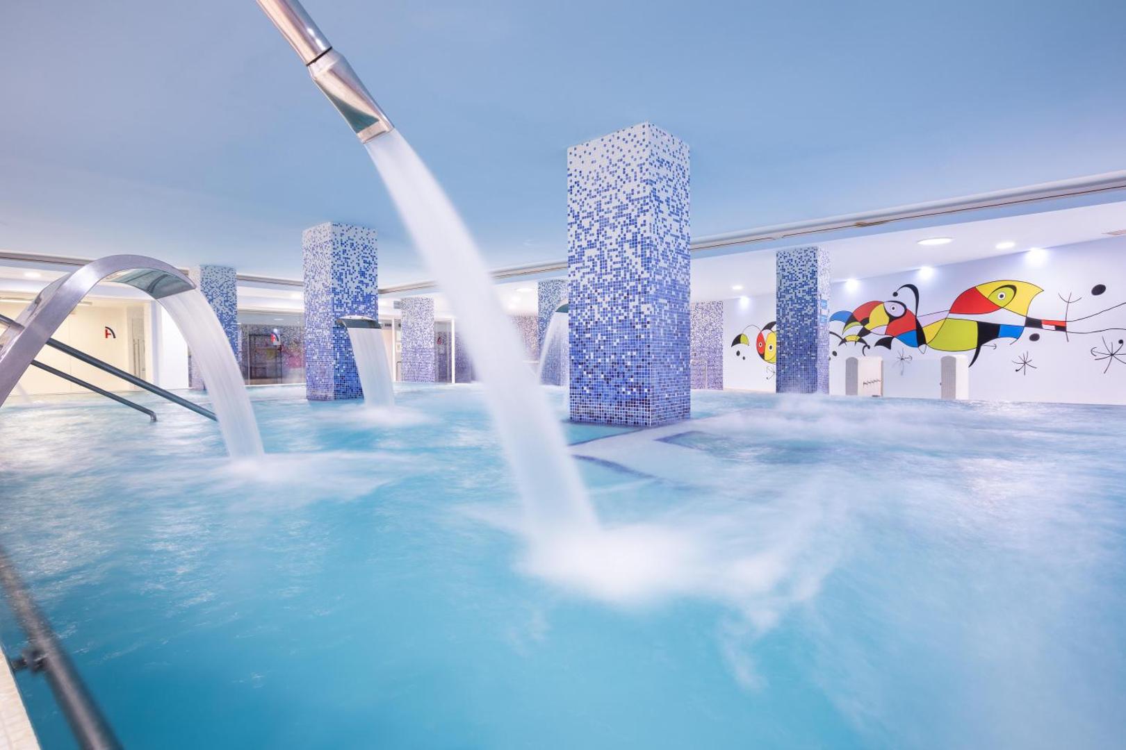 Alexandre Hotel Gala iekštelpu spa baseins ar kaskādēm un masāžas strūklām — gaiša atpūtas zona ar ziliem mozaīkas pīlāriem