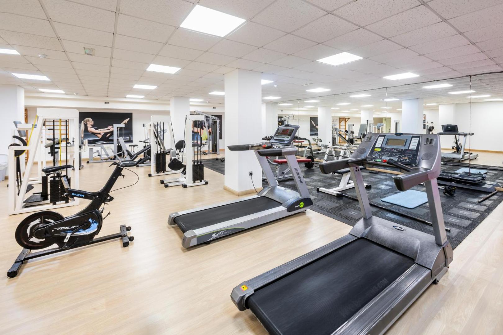 Alexandre Hotel Gala plaša fitness zāle ar moderniem skrejceliņiem, velotrenažieriem un spēka trenažieriem gaišā telpā ar spoguļiem