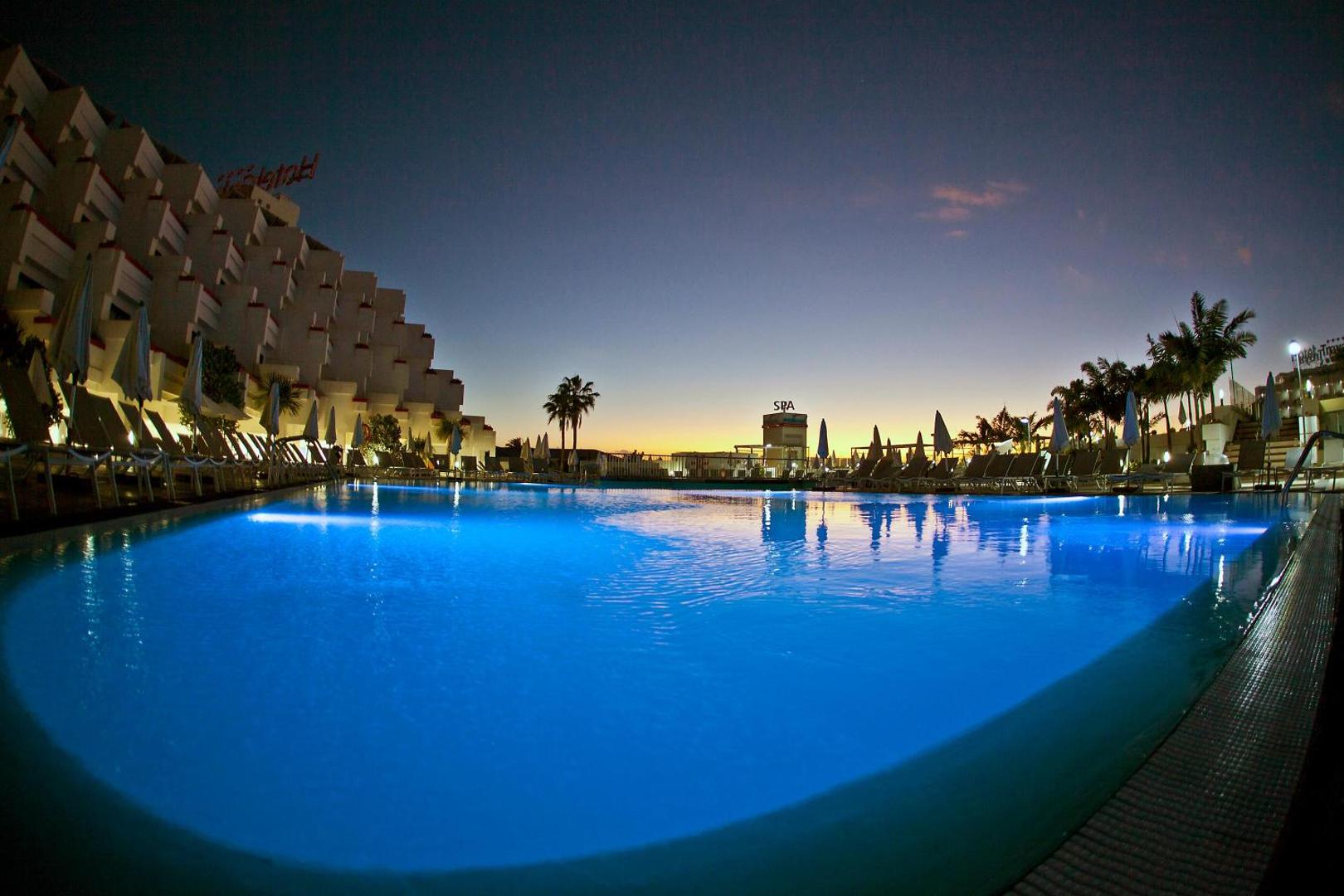 Alexandre Hotel Gala āra baseins vakarā Tenerifē — izgaismots zils ūdens, sauļošanās krēsli un viesnīcas fasāde pret saulrieta debesīm