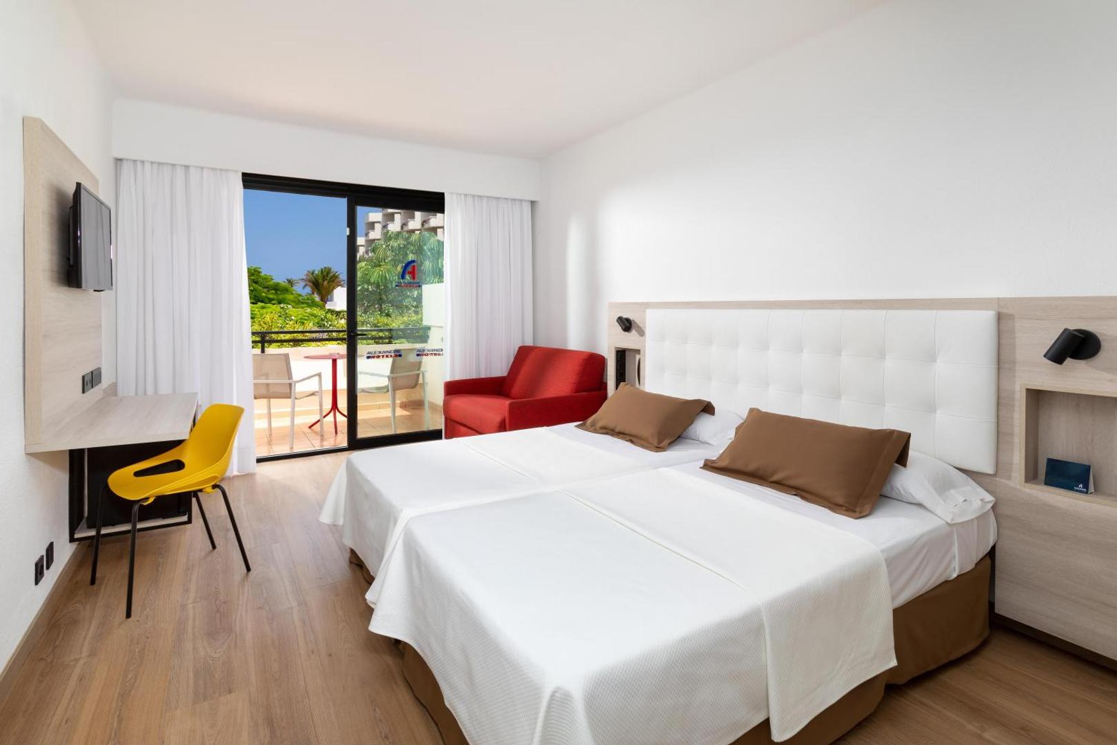 Gaišs Alexandre Hotel Gala viesnīcas numurs ar divguļamo gultu, dzeltenu krēslu un izeju uz saulainu terasi Tenerifē