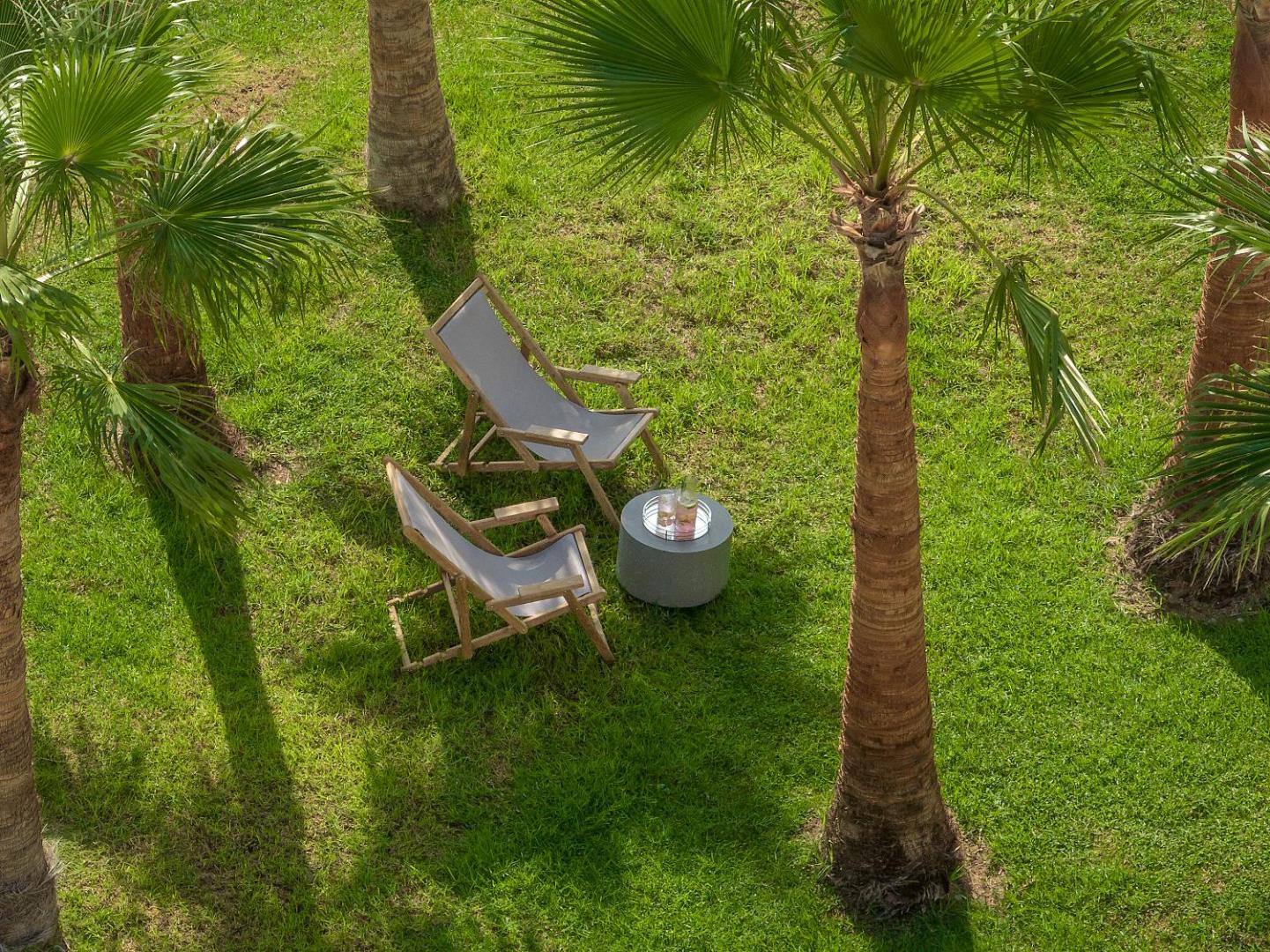 Grecotel Lux Me Dama Dama atpūtas zona dārzā ar koka zviļņiem un galdiņu zem palmām zaļā zālienā Rodas salā