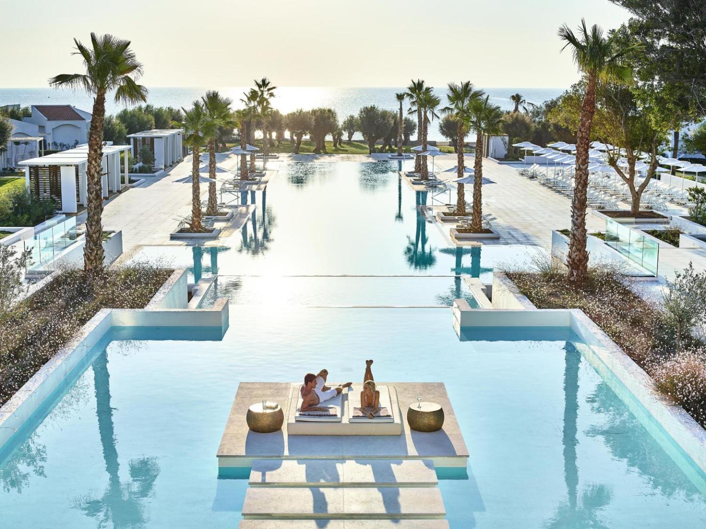 Grecotel Lux Me Dama Dama lielais āra baseins ar palmām Rodā — pāris atpūšas uz sauļošanās platformas ūdens vidū saulrietā