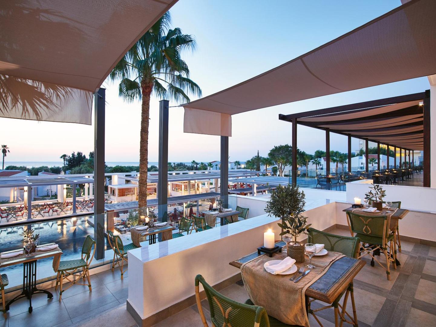 Grecotel Lux Me Dama Dama restorāna terase vakarā ar skatu uz jūru, izgaismotiem galdiņiem un kūrorta teritoriju
