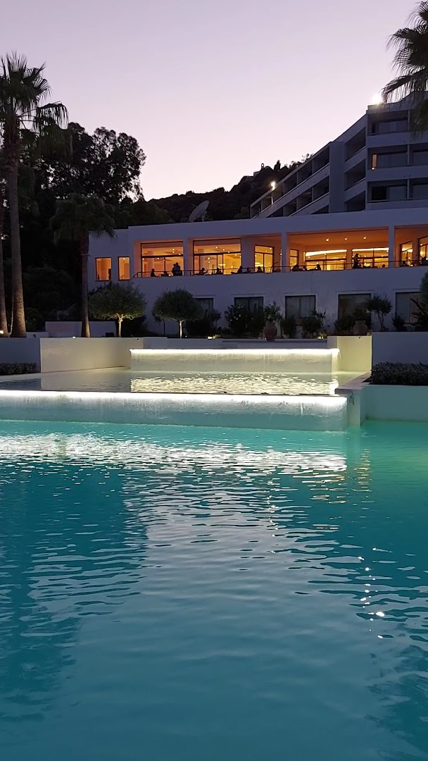 Grecotel Lux Me Dama Dama izgaismots baseins ar kaskādi vakarā, fonā redzama viesnīcas ēka un restorāna zona Rodā