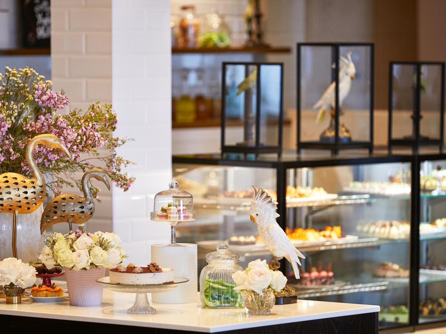 Grecotel Lux Me Dama Dama konditorejas nodaļa ar kūkām, ziediem un dekoratīvām figūrām — eleganta desertu bufete viesnīcas restorānā