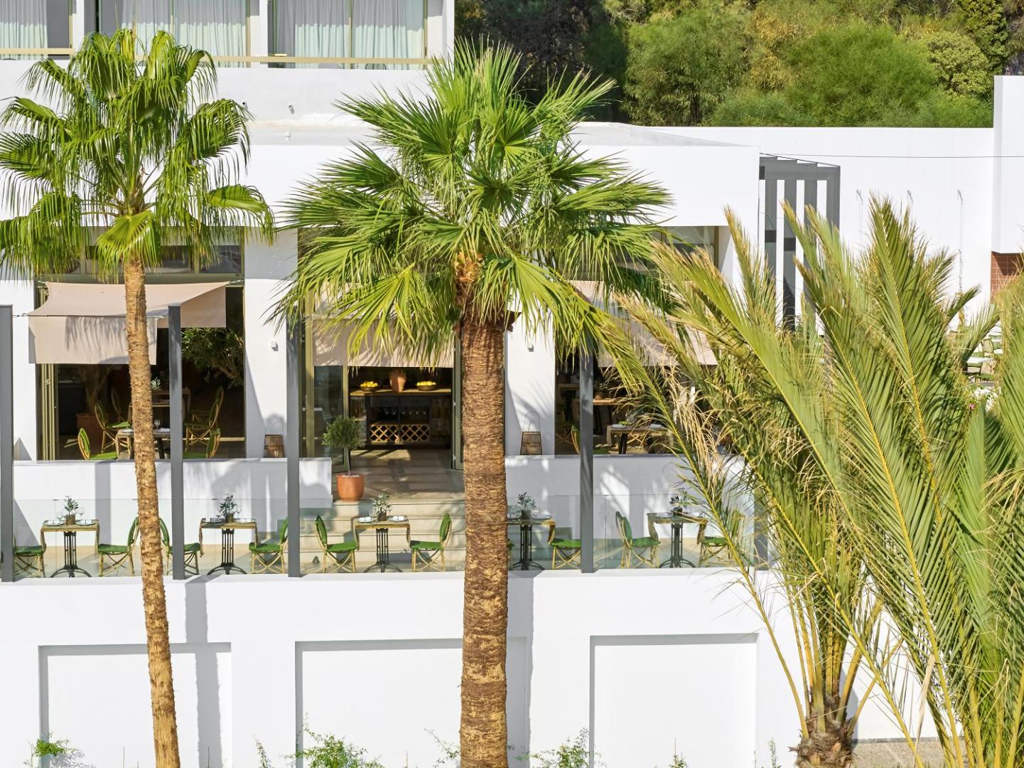 Grecotel Lux Me Dama Dama viesnīcas restorāna āra terase Rodas salā ar galdiņiem, zaļiem krēsliem un palmām saulainā dienā