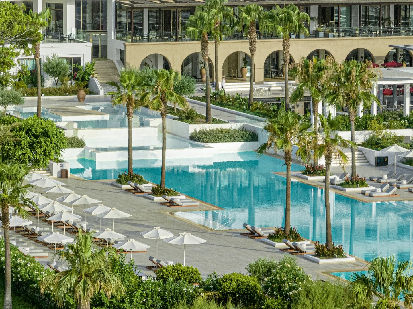 Grecotel Lux Me Dama Dama plašā baseinu zona Rodas salā ar palmām, sauļošanās krēsliem un modernu viesnīcas arhitektūru fonā