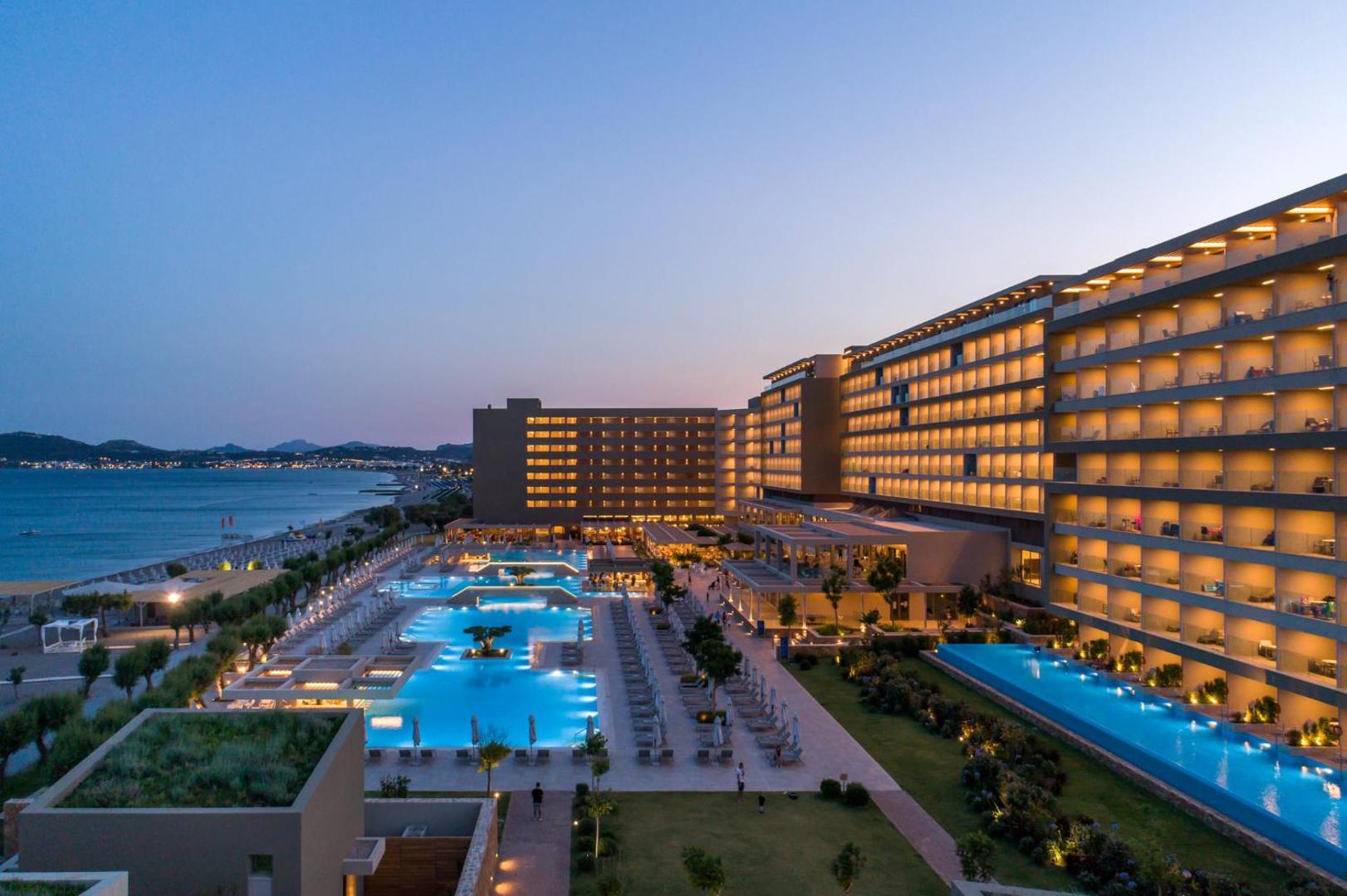 Amada Colossos Resort panorāmas skats vakarā — izgaismoti baseini, viesnīcas korpuss un Rodas piekrastes līnija saulrietā