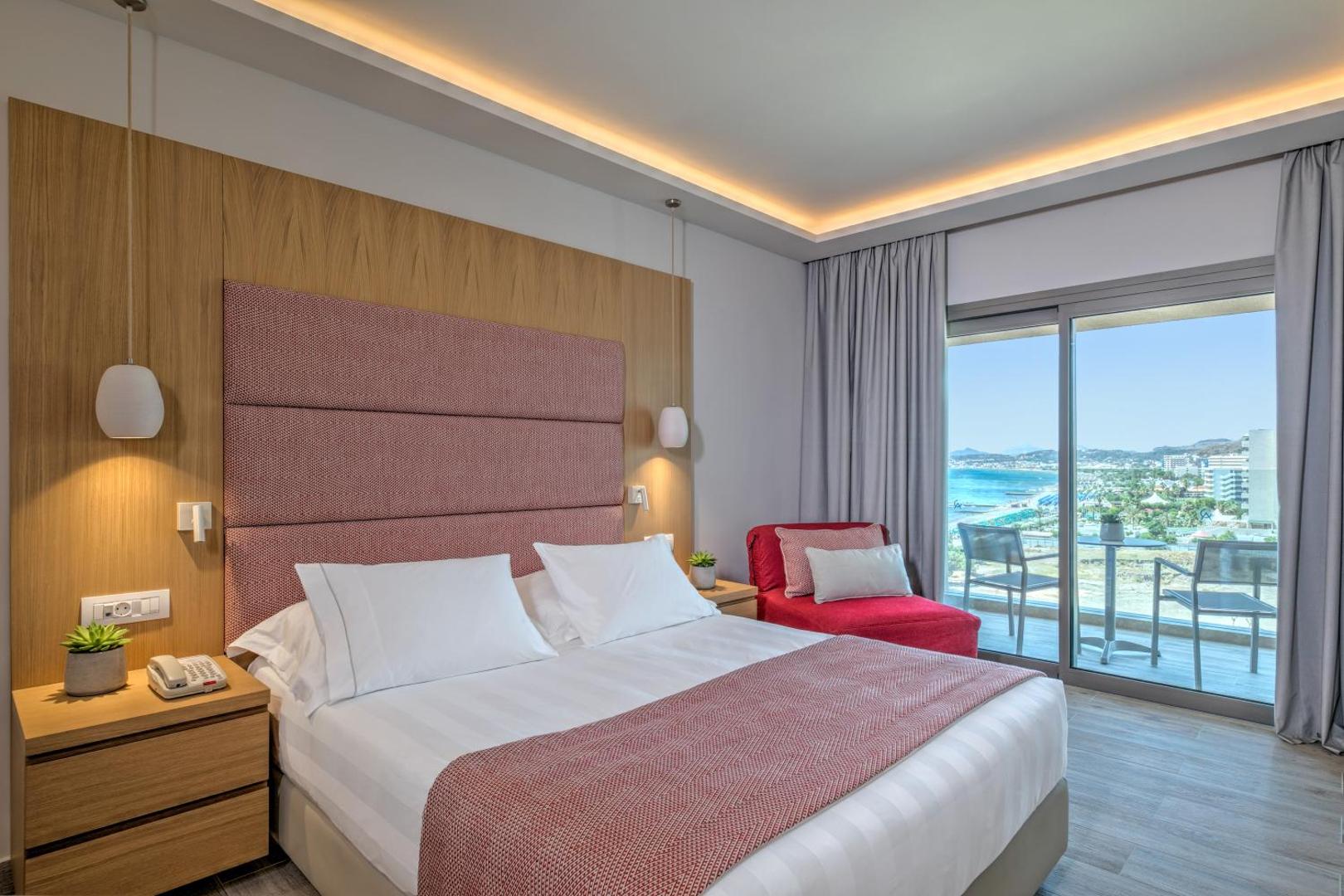 Amada Colossos Resort gaišs numurs ar divguļamo gultu, sarkanu atpūtas krēslu un balkonu, no kura paveras skats uz piekrasti