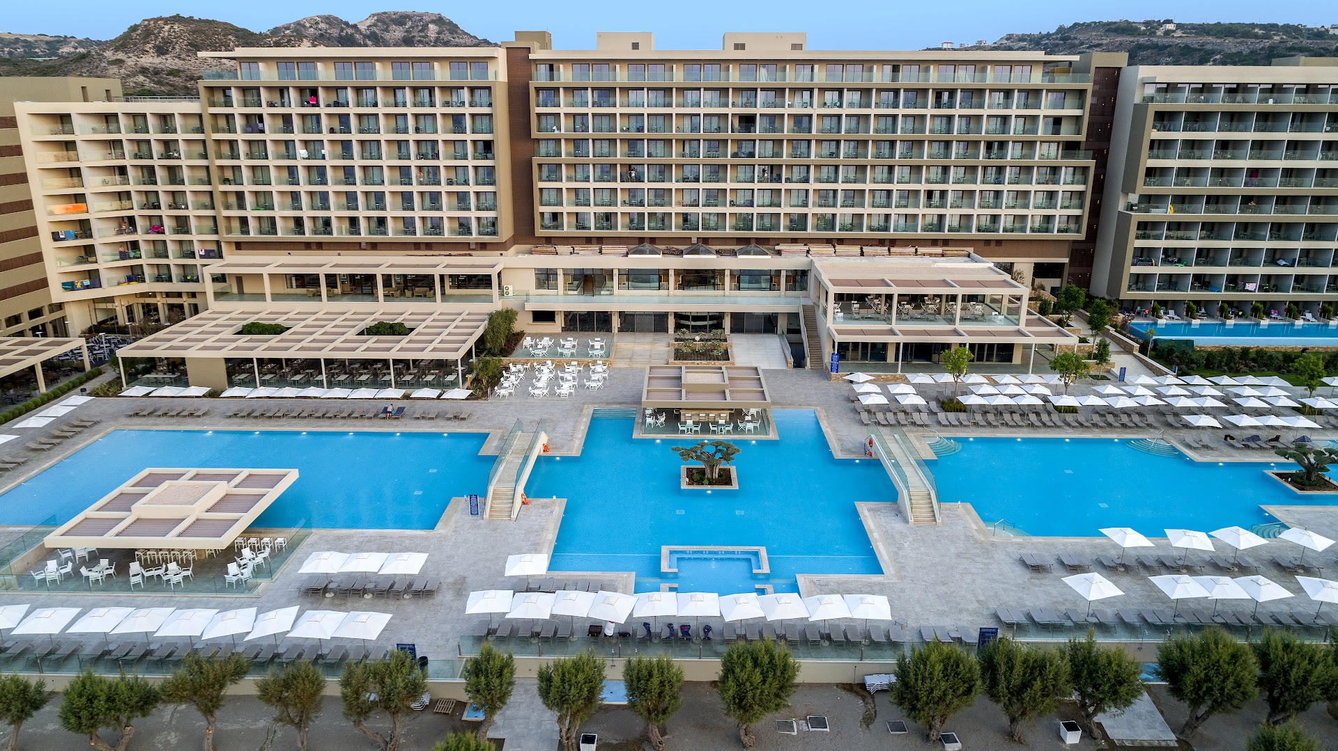 Amada Colossos Resort centrālā baseinu zona ar sauļošanās krēsliem un galveno ēku dienas laikā Rodā