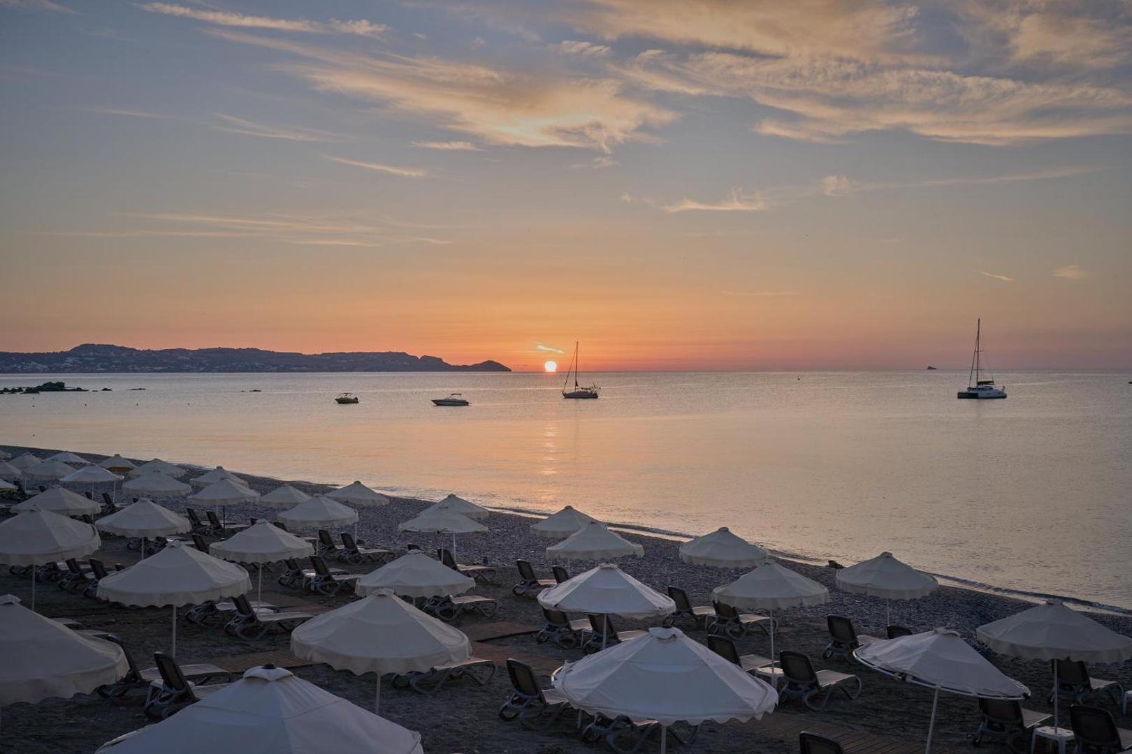 Saulriets Lindos Imperial Resort & SPA pludmalē — balti saulessargi priekšplānā un jahtas jūrā uz oranžu debesu fona