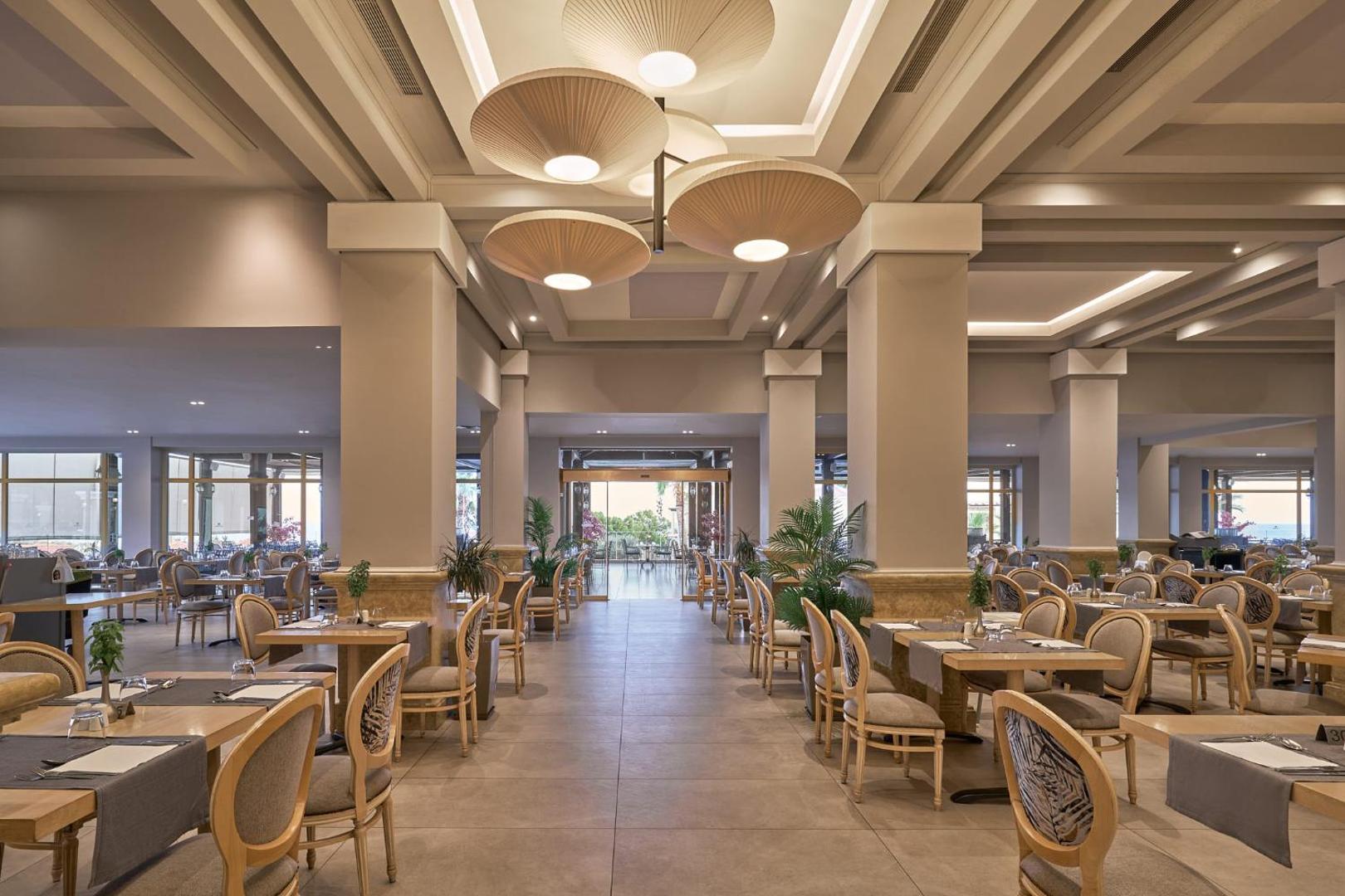 Lindos Imperial Resort & SPA galvenā restorāna iekštelpas ar elegantiem galdiem, krēsliem un modernu apgaismojumu
