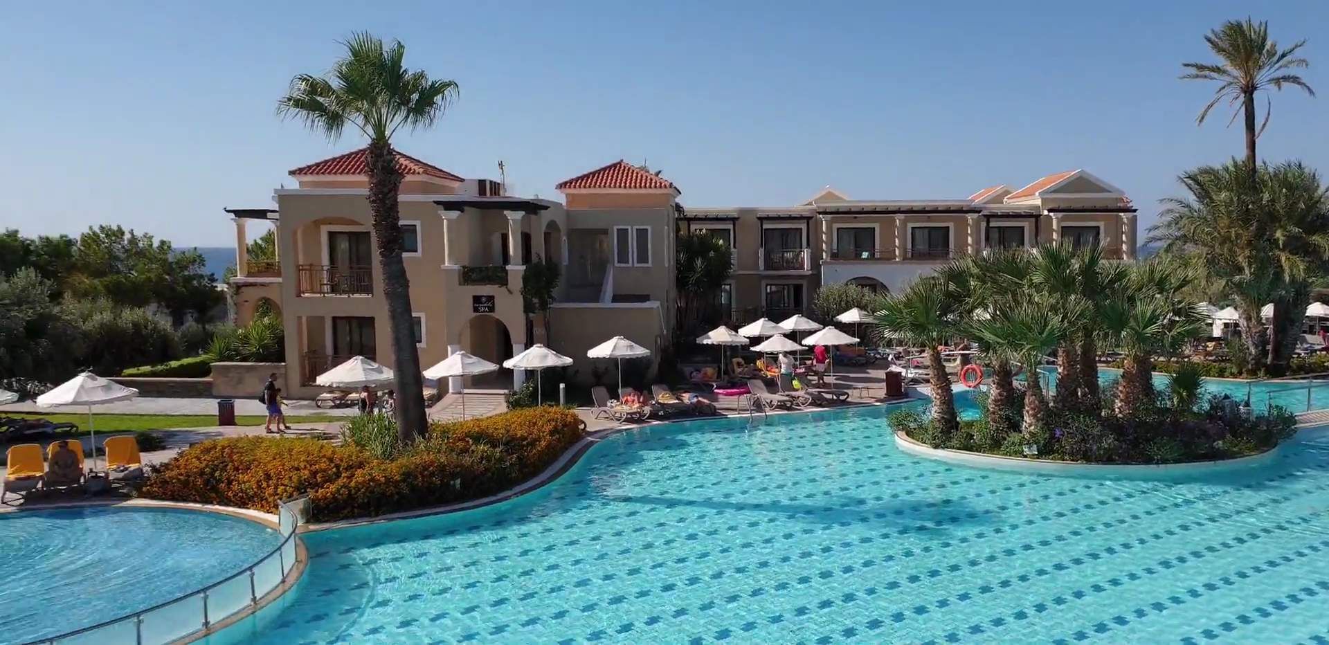 Lindos Imperial Resort & SPA atpūtas zona pie baseina ar palmām un saulessargiem — skats uz divstāvu numuru korpusiem Rodas salā