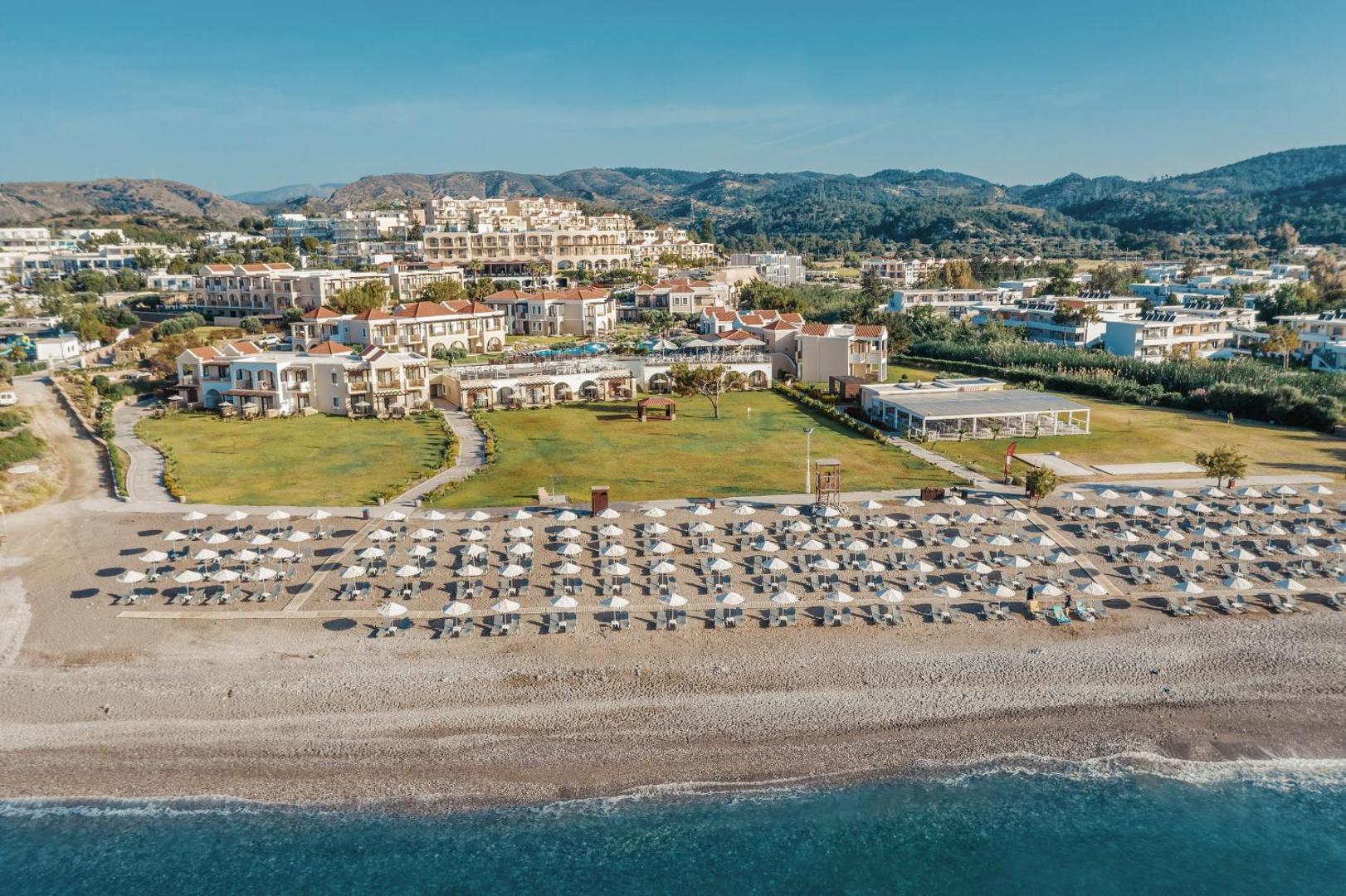 Lindos Imperial Resort & SPA panorāmas skats no putna lidojuma Rodas salā — viesnīcas komplekss pie plašas oļu pludmales ar kalniem fonā